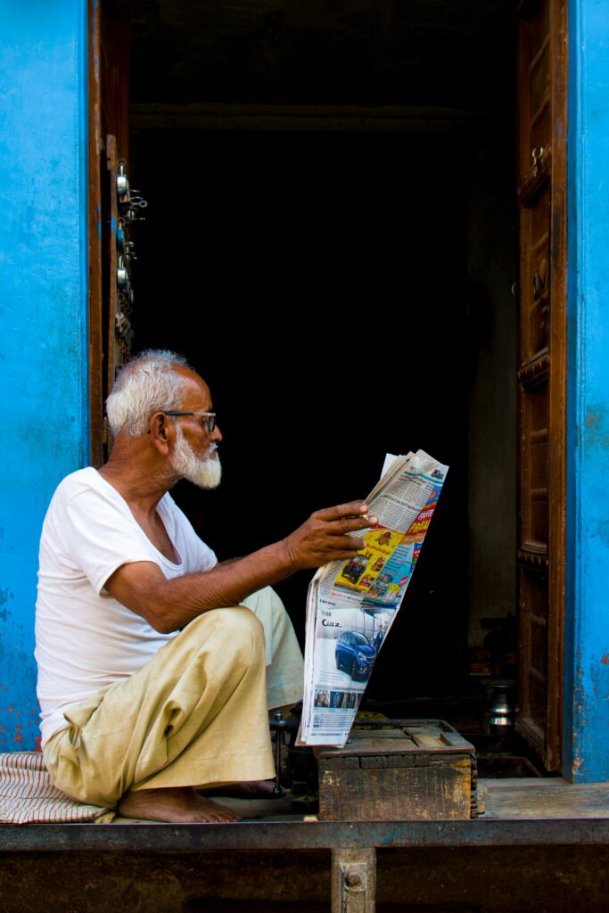 pexels-photo-2248503-2248503 Senior man in white shirt reading a newspaper in a vibrant blue doorway in Bikaner, India.